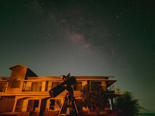 A reflector telescope on a tripod in front of a two-storey building at night, with stars and the Milky Way overhead.